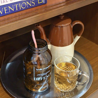 A styled tea setup on a round metal tray, featuring a dark amber glass jar with a pink straw and a visible label reading 'web organic DEFENCE', a clear glass mug filled with tea, and a brown-and-cream ceramic teapot in the background.