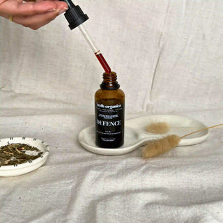 Brown glass bottle with dropper labeled 'Defence' by Welb Organics on a white textured surface next to dried herbs on a small plate.