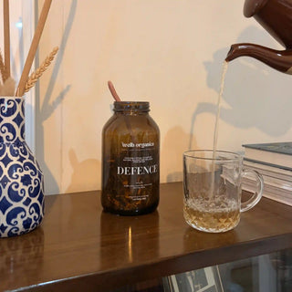 A dark amber glass jar labeled 'health organiser DEFENCE' sits on a wooden table with tea being poured from a brown teapot into a clear glass mug. A blue-and-white patterned vase with dried wheat is visible on the left.