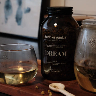 Brown jar of herbal tea labelled "Welb Organics Dream" on a wooden tray next to a glass and a spoon with dried chamomile.