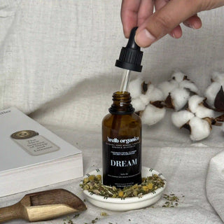 Brown bottle of herbal tincture with a label "Welb Organics Dream" on a small plate with dried herbs, next to a branch of cotton, a book and a wooden spoon.