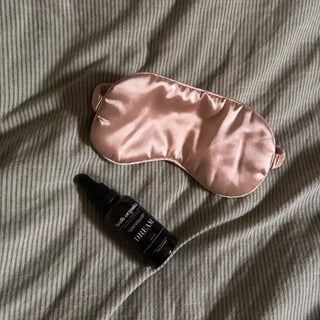 A pink satin sleep mask and a small black dropper bottle labeled 'DREAM' resting on a gray ribbed fabric background.