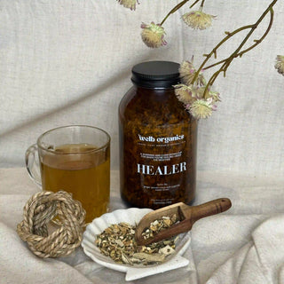 Brown jar of herbal tea labelled "Healer" by Welb Organics on a textured surface next to a tea cup and dried herbs on a small plate with a wooden spoon.