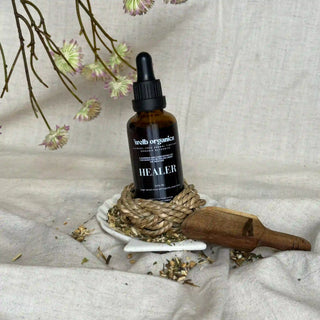 Bottle of herbal tincture labeled 'Healer' on a woven knot with dried herbs and wooden spoon on a textured fabric background