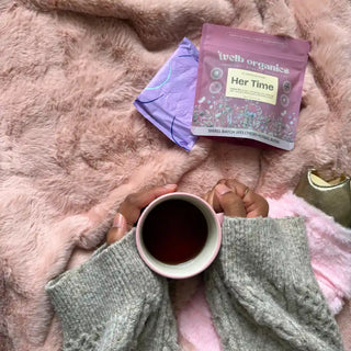A person holds a mug of tea wrapped in a grey knit sweater, lying on a fluffy pink blanket next to a packet of Welb Organics Her Time herbal tea.