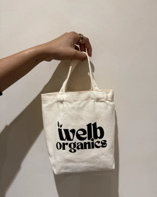 White tote bag with 'welb organics' text held by a hand against a plain background