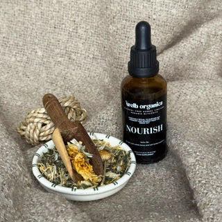 Bottle of herbal tincture labeled 'Nourish' next to a bowl of dried herbs on a textured surface