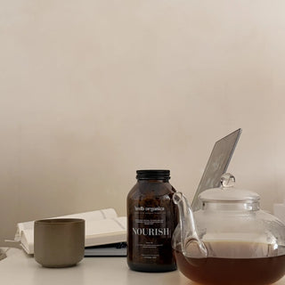 A dark amber bottle labeled 'nourish' by 'health organics' sits on a light-colored tabletop next to a clear glass teapot filled with tea, an open notebook, and a small gray cup against a plain beige wall.