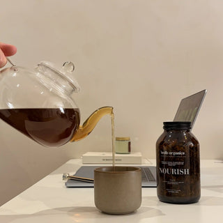 A hand holds a clear glass teapot pouring dark coffee into a matte taupe ceramic cup on a white table. A brown amber bottle labeled 'nourish' stands nearby with a laptop and books in the background.