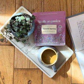 A package of Welb Organics Second Spring herbal tea is displayed on a white tray alongside a small potted plant and a cup of coffee on a wooden floor.