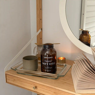 A dark amber glass jar abeled 'SOFT DAYS' by 'welb organics' sits on a glass tray on a light wood desk, styled with a grey ceramic mug, a gold candle, and an open book in a warm, minimalist room setting.