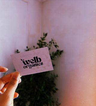 Hand holding a card saying 'Welb Organics' with a bplant and wall in the background